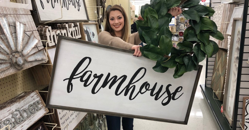 sara holding a farmhouse sign and magnolia wreath in the store aisle 