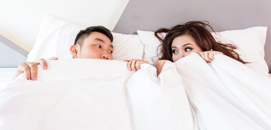 man and woman under white comforter in bed bedding looking at each other