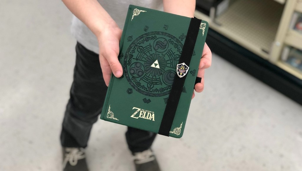 hands holding a dark green and gold zelda writing journal with shoes in background