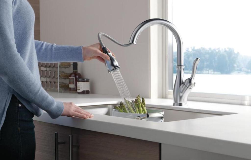 woman wearing blue shirt holding spray head on stainless faucet rinsing off vegetables 