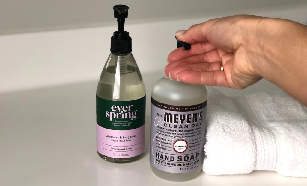 hand pushing down on lavender mrs meyers hand soap next to bottle of purple everspring on white countertop