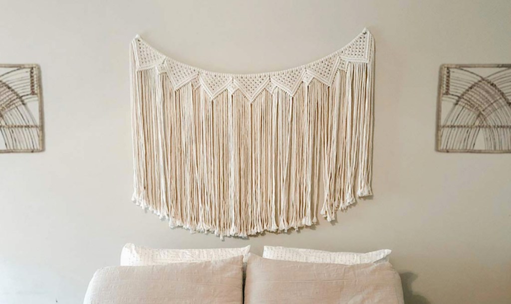 cream colored macrame wall hanging banner on gray wall with bed and pillows below
