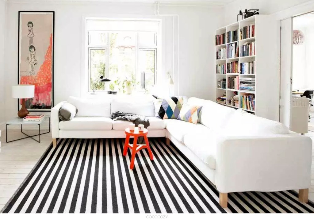 black and white stripe rug in living room with large white sectional couch and red stool and bookshelves